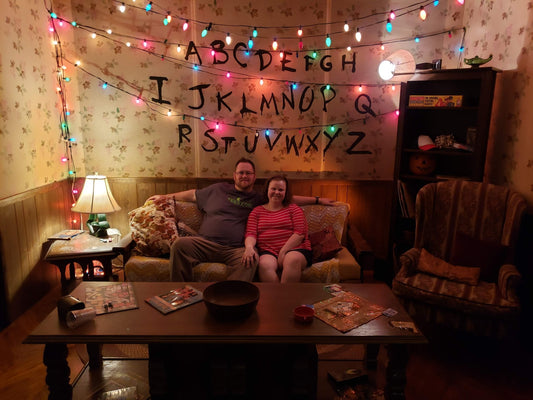Jeff and Sam sitting on the couch in front of Joyce Byers' alphabet wall with Christmas lights, a throwback from their first Halloween Horror Nights